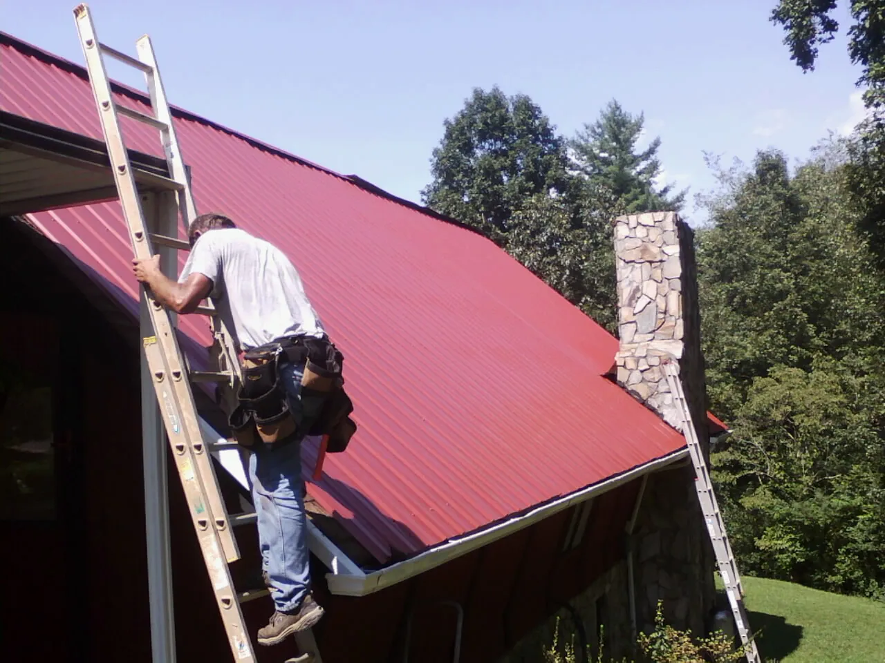 Roofer on a ladder beside a red metal roof and stone chimney — Soffit & Fascia Repair by Roofing Brothers in Underwood